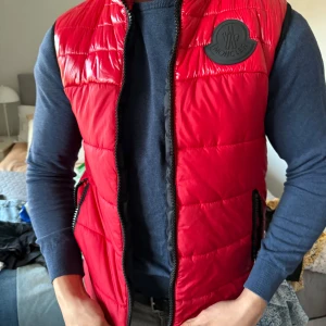 Röd dunväst från Moncler - Snygg röd dunväst från Moncler med svart logotyp på bröstet och dragkedja framtill. Västen har en quiltad design och två fickor med dragkedja. Perfekt för lager-på-lager-stil.