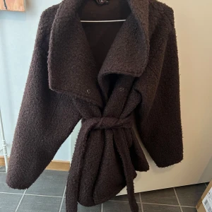 Mörkbrun teddyjacka med bälte - Mörkbrun teddyjacka med stor krage och bälte i midjan. Jackan har lång ärm och en oversized passform. Säljes pga fick en annan färg i present. Strl M