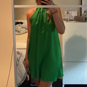 Grön halterneckklänning klänning satin från Dixie - Säljer en grön halterneckklänning från Dixie i glansigt material. Klänningen har en lös passform, är ärmlös och knyts i nacken med en rosett. Perfekt för sommarens tillfällen när du vill sticka ut med färg.