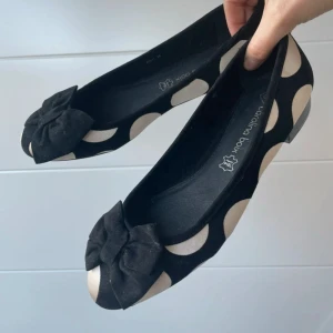 Svarta ballerinaskor med stora vita prickar från Carolina Boix - Säljer ett par svarta ballerinaskor från Carolina Boix med stora vita prickar och en söt rosett framtill. Skorna har platt sula och rund tå, perfekta för dig som gillar lekfull stil. Materialet är textil.