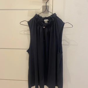 Svart ärmlös blus med volangkrage - Säljer en svart ärmlös blus med hög volangkrage och liten knappdetalj framtill. Blusen har en rak passform och är tillverkad i ett mjukt, lätt material som faller snyggt.