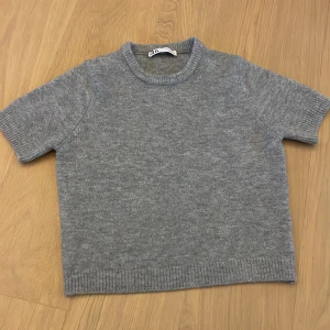 Grå stickad tröja från Zara - Mysig grå stickad tröja från Zara med korta ärmar och rund halsringning. Tröjan har ribbade muddar vid ärmslut och nederkant, perfekt för lager-på-lager-stil. Enkel och stilren design som passar till mycket. Inte använd mycket och skulle säga att den passar xs också eftersom jag har xs men den passar perfekt på mig. Skriv för bilder när jag har den på mig.