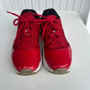 Röda Jordan 11 - Säljer ett par röda Jordan sneakers med vita detaljer och svart insida. Skorna har snörning, rund tå och platt sula. Ovandelen är i skinn och syntet, med vit Jumpman-logga på sidan. Perfekta för dig som gillar sportig stil.