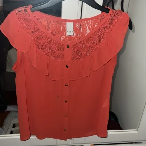 Korallröd blus med spets och volang från Vela - Säljer en korallröd blus från Vela med vacker spets upptill och volangdetaljer längs axlar och bröst. Blusen har knappar framtill och är ärmlös, perfekt för en färgstark look. Aldrig använd
