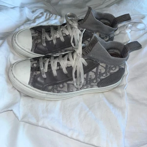 Grå höga sneakers från Christian Dior - Säljer ett par grå höga sneakers från Christian Dior med tydlig logotyp på sidan och tungan. Skorna har snörning, vit sula och detaljer i både textil och syntetmaterial. Perfekta skor som funkar till jeans, byxor och kjol! Köpta för mindre än ett år sedan! En intressekoll då jag inte är säker på att jag vill sälja! Skriv om det finns några frågor💞