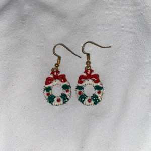 Julkrans-örhängen med rosett - Säljer ett par örhängen formade som julkransar med gröna och röda detaljer samt en röd rosett upptill. Perfekta för att sprida julkänsla! Hängande modell med guldfärgade krokar.