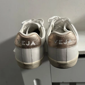 Vita sneakers från Veja med guldiga detaljer - Säljer ett par snygga vita sneakers från Veja med metallicguld detaljer på häl och sidor. Skorna har klassisk låg modell, rund tå och vita skosnören. Perfekta för en stilren vardagslook.