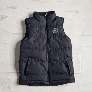 Svart väst Warp - Säljer en svart dunväst från Warp med hög krage och dragkedja framtill. Västen har två fickor med dragkedja och en logotyp på bröstet. Perfekt för lager-på-lager under kyliga dagar. Priset är inte hugget i sten, öppen för förslag.