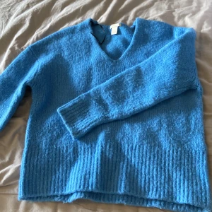 Blå stickad tröja med v-ringning - Säljer en blå stickad tröja med v-ringad hals. Tröjan har lång ärm och ribbade muddar vid ärmslut och nederkant. Perfekt för kyligare dagar och enkel att matcha med olika outfits.