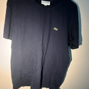 Mörkblå t-shirt från Lacoste - Säljer en klassisk mörkblå t-shirt från Lacoste med den ikoniska gröna krokodilloggan broderad på bröstet. T-shirten har rund hals och är tillverkad i mjuk bomull. Perfekt basic-plagg för garderoben.