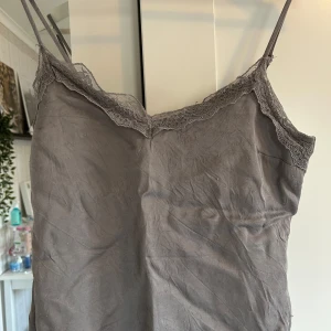 Grå linnetopp med spets från Kappahl - Säljer en grå linnetopp från Kappahl med tunna axelband och vacker spetsdetalj längs urringningen. Perfekt för lager-på-lager eller sommarens varma dagar. Tunn och mjuk i materialet. Toppen är lite nopprig.