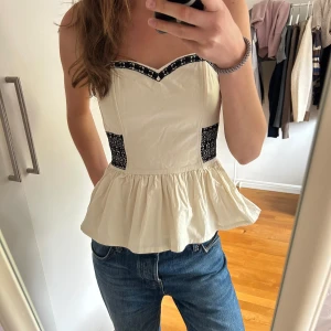 Vit korsett-topp med svarta detaljer - Jättesöt vit (krämvit) korsett-topp (ursprungligen en klänning men sytt om den till topp). Finns en dragkedja på ena sidan. Baktill har den smock vilket gör att den passar flera storlekar, jag själv är S och den passar bra.