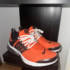 Nike Presto sneakers i orange och svart - Säljer ett par Nike Presto sneakers i en snygg orange färg med svarta och vita detaljer. Skorna har vit sula, svart Nike-logga och vita snören. Perfekta för dig som vill sticka ut med färg på fötterna.