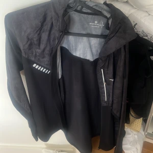 Svart vindjacka från SOC - Svart vindjacka med huva från SOC. Jackan har reflekterande detaljer på bröstet och en dragkedja framtill samt på bröstfickan. Lätt material och lång ärm, perfekt för utomhusaktiviteter.