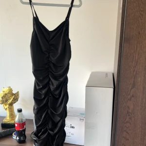 Svart omlottklänning med rynkade detaljer - Säljer en svart långklänning med smala axelband och snygga rynkade detaljer längs hela framsidan. Klänningen har en figurnära passform och är perfekt för dig som vill sticka ut. Materialet känns mjukt och följsamt.