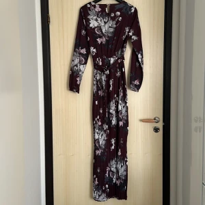 Blommig långklänning från Lorraine - Säljer en vinröd långklänning från Lorraine med vackert blommigt mönster i grått och vitt. Klänningen har långa ärmar, knytband i midjan och plisserad kjol. Perfekt för dig som gillar romantisk stil och vill sticka ut.