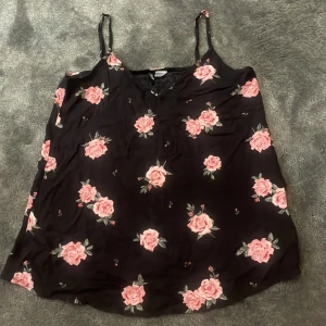 Svart blommig linnetopp med rosa rosor - Säljer en svart linnetopp med tunna axelband och blommigt mönster av rosa rosor och gröna blad. Perfekt för varma dagar och enkel att matcha med jeans eller kjol.