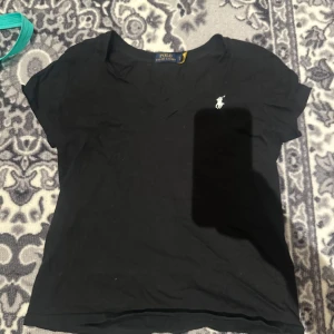 Svart t-shirt från Polo Ralph Lauren - Svart t-shirt från Polo Ralph Lauren med klassisk v-ringning och vit broderad logga på bröstet. T-shirten har korta ärmar och är tillverkad i mjukt material som passar perfekt till vardags.