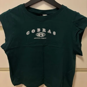 Mörkgrön croppad t-shirt med tryck 'Cobras Arizona Cobras' - Säljer en mörkgrön croppad t-shirt med vitt tryck 'Cobras Arizona Cobras' framtill. T-shirten har rund halsringning och korta ärmar. Perfekt för en avslappnad stil.