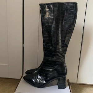 Svarta stövlar med krokodilmönster - Säljer ett par svarta höga stövlar med glansig krokodilpräglad yta och bred klack. Stövlarna har spetsig tå och dragkedja på insidan. Perfekta för dig som vill sticka ut med en trendig look. De är väldigt bekväma och är storlek 39 men passar mer som storlek 38. 