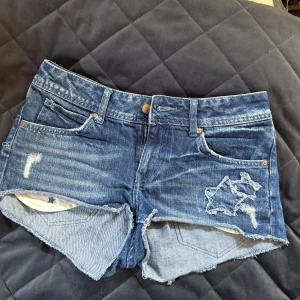 Blå jeansshorts  - Snygga blå jeansshorts med slitningar och en  stjärnformad detalj på ena benet. Passar mer en storlek 36 än 38 