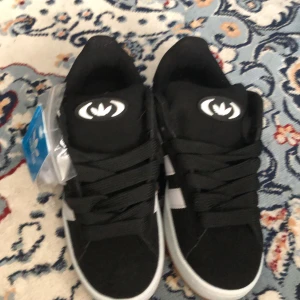 Svarta Adidas sneakers med vita ränder - Säljer ett par klassiska svarta Adidas sneakers med vita ränder på sidorna och vit sula. Skorna har svart snörning och gummisula i brunt. Perfekta för en sportig och stilren look. Extra vita skosnören medföljer.