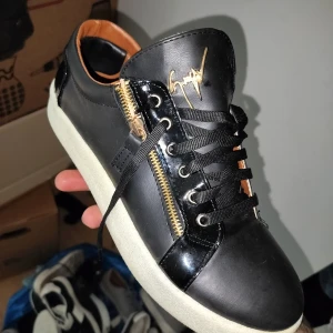 Svarta sneakers från Giuseppe Zanotti med gulddetaljer - Säljer ett par svarta sneakers från Giuseppe Zanotti med vita sulor, guldfärgade dragkedjor på sidorna och svart snörning. Skorna har en lyxig känsla med läderdetaljer och signatur-logga på plösen.