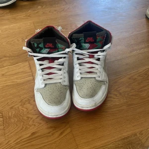 Nike Air Jordan 1 Retro High OG - Säljer ett par Nike Air Jordan 1 Retro High OG med vit och beige mocka, röda detaljer och vit sula med röd undersida. Skorna har snörning och ikonisk Air Jordan-logga på sidan. Insidan har färgglatt mönster och ovandelen är i mix av skinn och mocka.