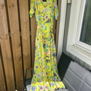 Gul blommig långklänning med knytband - Säljer en somrig gul långklänning med blommigt mönster och v-ringning. Klänningen har korta ärmar och knytband i midjan för en fin passform. Perfekt för varma dagar och midsommarfirande.