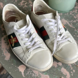  vita sneakers med broderad bi - Säljer ett par vita sneakers med klassiska gröna och röda ränder på sidorna och en broderad guldfärgad bi. Baksidan har metallicröd detalj på ena skon och metallicgrön på den andra. Skorna har vit snörning och rund tå.