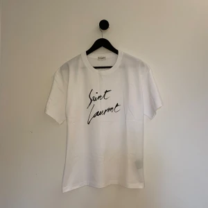 Saint Laurent T-shirt - Säljer en vit t-shirt från Saint Laurent med svart handskriven logga på bröstet. Klassisk passform och rund halsringning. Perfekt för dig som gillar stilrena och exklusiva plagg. Kvitto o tags finns, använd sparsam och mycket kvar att ge! Storlek M fast kan även passa S