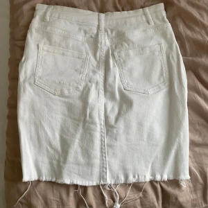 Vit jeanskjol med rå kant - Vit jeanskjol med klassiska femficksdetaljer och rå, fransig nederkant. Kjolen har dragkedja och knapp framtill samt bälteshällor i midjan. Perfekt för en avslappnad och trendig look. Storlek 36