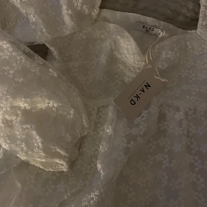 Studentklänning/Vit blommig klänning från NA-KD  - Superfin vit klänning från NA-KD med blommigt mönster. Klänningen har långa puffärmar, markerad midja och söt sweetheart-ringning. Perfekt för vår och sommar. Även perfekt för studenten!!! 