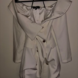 Vit omlottklänning med stor krage, kavajklänning - Säljer en vit kortklänning med stor, dramatisk krage och omlott-design. Klänningen har långa ärmar, knappar framtill och fickor. Perfekt för dig som vill sticka ut med en unik och elegant look.