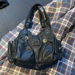 Svart axelväska - Säljer en svart axelväska med flera fack och dragkedjor. Väskan har två rejäla handtag, dekorativ och detaljer i silverfärg. Tillverkad i syntetmaterial med praktiska sidofickor och en tydlig logotyp framtill. Långt handtag kommer med så du kan ha den som crossbody