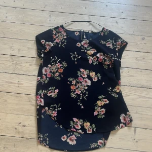 Mörkblå blommig blus - Säljer en mörkblå blus med färgglatt blommönster. Blusen har rund halsringning, korta ärmar och är lite längre baktill. Perfekt för dig som gillar en feminin och bekväm stil.