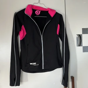 Svart träningsjacka med rosa detaljer från Woxer - Sportig svart träningsjacka från Woxer med dragkedja och rosa insida samt detaljer. Jackan har långa ärmar med vita sömmar och två fickor framtill. Perfekt för aktiva dagar och träning.