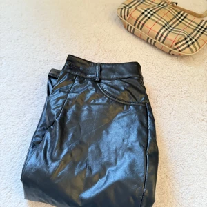 Svarta skinnbyxor - Snygga svarta byxor i skinnimitation med normal passform och klassisk femficksdesign. Perfekta för att lyfta vilken outfit som helst med en cool vibe. Byxorna har bälteshällor och stängs med dragkedja och knapp.