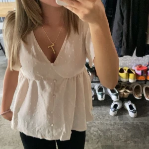 Beige blus med knappar och volang - Söt beige blus med kort ärm, knappar framtill och volang nedtill. Blusen har v-ringning och ett diskret prickigt mönster i tyget. Perfekt för en fräsch och somrig look.