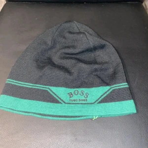 Svart och grön mössa från Hugo Boss - Snygg svart mössa med gröna detaljer och broderad Hugo Boss-logga framtill. Mössan har en sportig design med gröna ränder och är tillverkad i mjukt material. Perfekt för kyliga dagar. Storleken är one size och passar dem flesta. Det är bara att höra av sig vid frågor och funderingar 😁