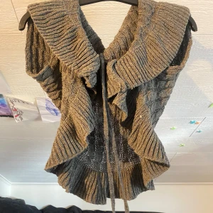 Grå stickad kofta med volangkant - Säljer en grå stickad kofta med bred volangkant och knytband framtill. Västen har ett snyggt kabelstickat mönster och stor krage som ger en cool och mysig look. Perfekt att slänga över en långärmad tröja för extra stil. 