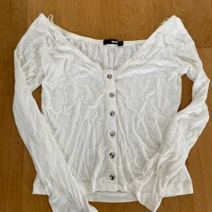 Vit ribbad långärmad topp från Bik Bok - Säljer en vit långärmad topp från Bik Bok med ribbad struktur och knappar framtill. Toppen har en v-ringad hals och är tillverkad i ett mjukt material som sitter bekvämt på kroppen.