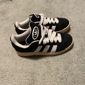 Svarta Adidas Campus sneakers med vita ränder - Säljer ett par klassiska Adidas Campus sneakers i svart mocka med vita ränder och vit sula. Skorna har breda vita skosnören och Adidas-logga på plösen. Perfekta för dig som gillar retrostil och streetwear.