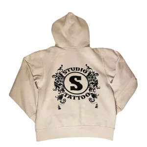 Studio Tattoo hoodie - Säljer en vit hoodie med dragkedja och huva. På ryggen finns ett stort svart tryck med texten 'Studio Tattoo' och dekorativa detaljer. Perfekt för dig som gillar streetwear och vill sticka ut lite extra.