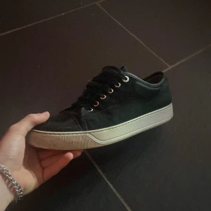 Svarta sneakers från Lanvin - Säljer ett par svarta sneakers från Lanvin med vita sulor och svarta skosnören. Skorna har rund tå och metallfärgade öljetter för snörningen. Klassisk och stilren design som passar till mycket.
