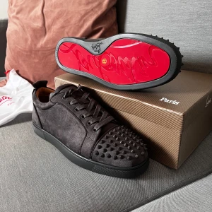 Svarta sneakers med nitar från Christian Louboutin - Säljer ett par svarta sneakers från Christian Louboutin i mocka med ikonisk röd sula och nitar framtill. Skorna har snörning och rund tå, samt en platt sula. Perfekta för dig som vill sticka ut med en lyxig och edgy stil.