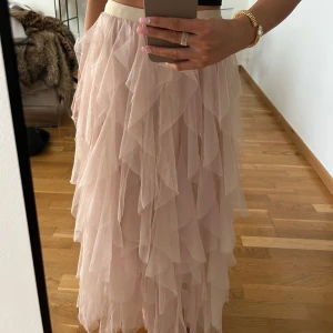 Ljusbeige/rosa volangkjol i tyll - Lång beige/ljusrosa kjol med flera lager volanger i tyll. Storlek XS. Sitter både bekvämt och fint på, som ny använt 1 gång. Plagget är i nyskick ✨