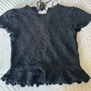 Svart spetsblus med volang - Säljer en svart blus i spets med korta ärmar och volang nertill. Blusen har knytning i nacken och ett fint mönster i spetsen. Aldrig använd