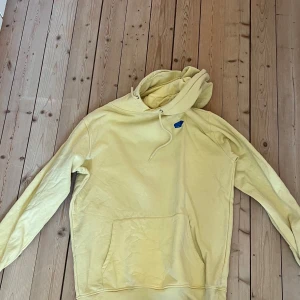 Gul hoodie med tryck från Air Cushion - Säljer en ljusgul hoodie från Air Cushion med stor svart och rosa logga på ryggen och liten blå detalj på bröstet. Tröjan har huva och magficka, perfekt för en avslappnad stil.