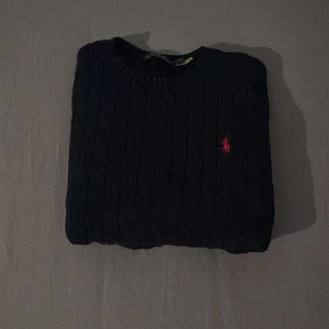 Svart kabelstickad tröja från Polo Ralph Lauren - Kabelstickad svart tröja från Polo Ralph Lauren med rund halsringning och klassisk röd broderad logga på bröstet. Tröjan har långa ärmar och ribbade muddar vid ärmslut och nederkant. Perfekt för lager-på-lager-stil.